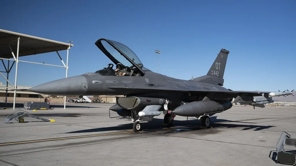 Grey USAF F-16 fighter jet with open canopy parked on tarmac under a clear blue sky, tail code OT visible.
