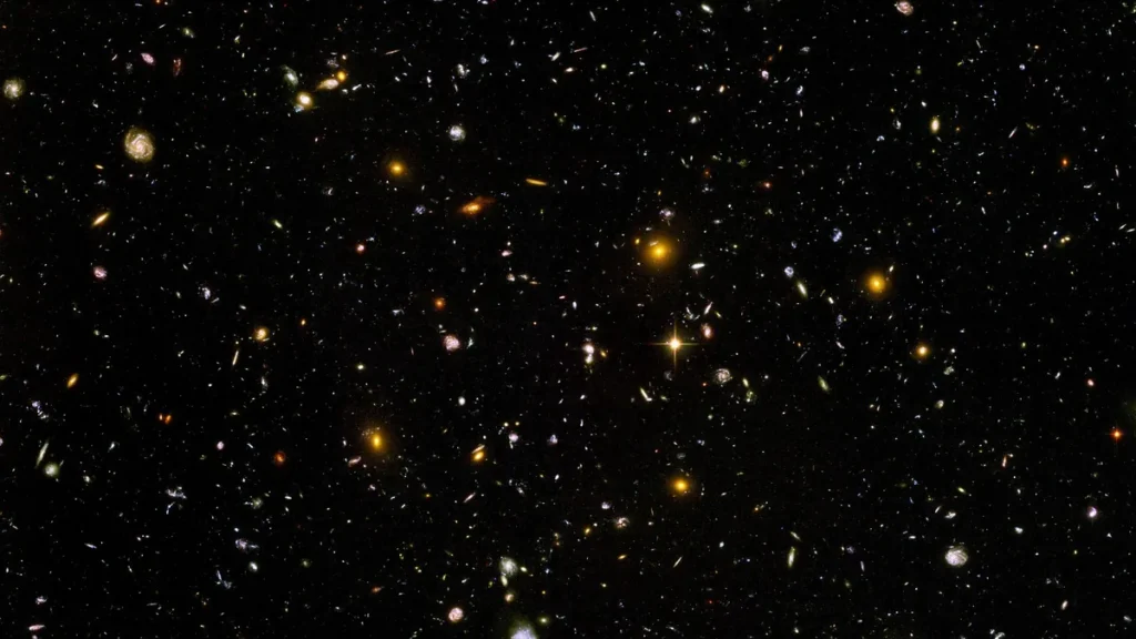 Deep field image showing thousands of colorful distant galaxies and a bright star against the black cosmic background.