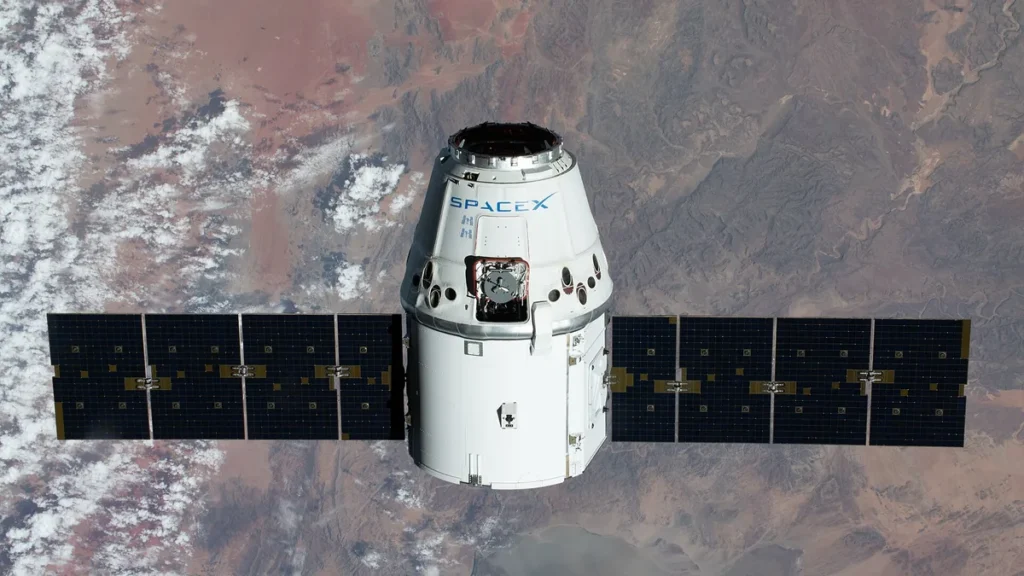 SpaceX Dragon capsule orbiting Earth with solar panels deployed against a desert landscape background.