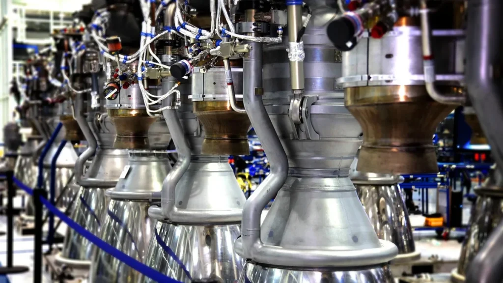 Row of metallic rocket engines with copper-colored nozzles and complex wiring in an assembly facility.