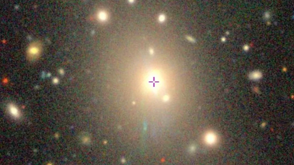 A colorful cosmic view of a galaxy cluster, with a purple crosshair marking the central bright galaxy.