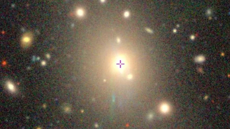 A colorful cosmic view of a galaxy cluster, with a purple crosshair marking the central bright galaxy.
