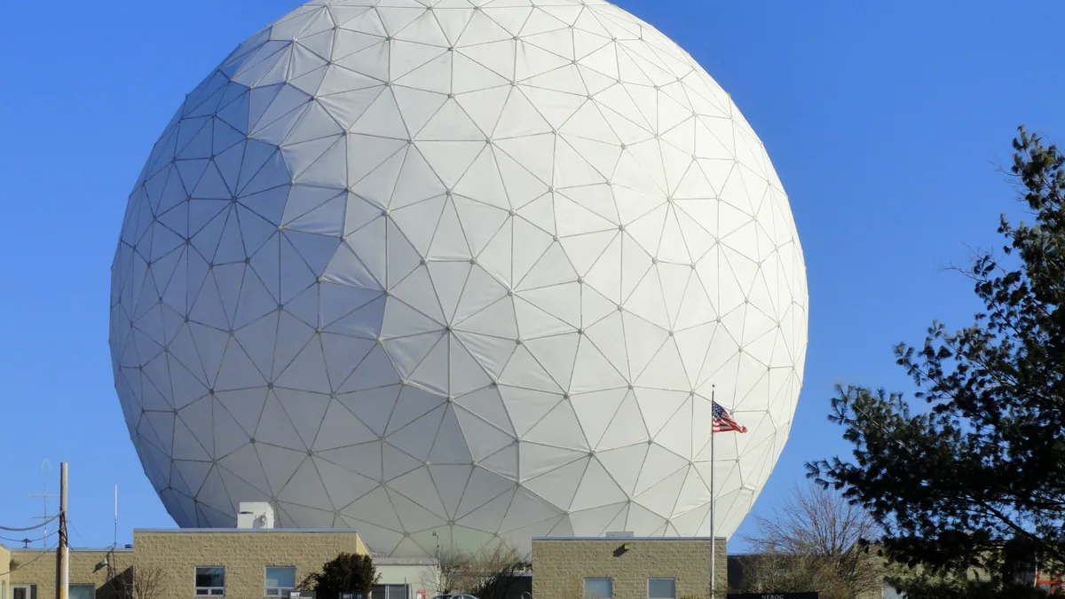 A giant white geodesic dome of a radio telescope stands against a clear blue sky, with a US flag flying in front.