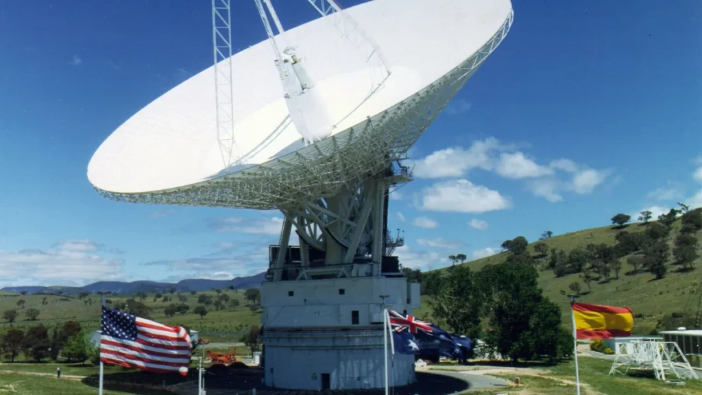 Large white satellite dish antenna with US, Australian, and Spanish flags under a bright blue sky.