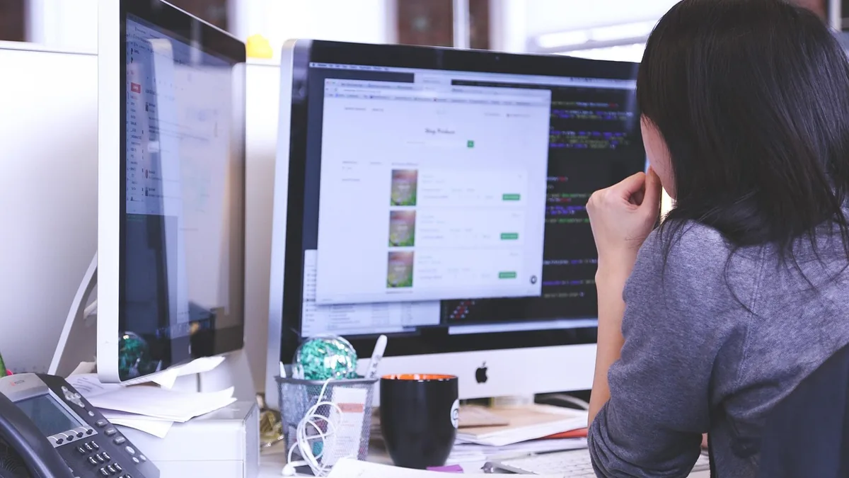 A woman viewed from behind works on a web layout and code on dual monitors at a tidy desk.