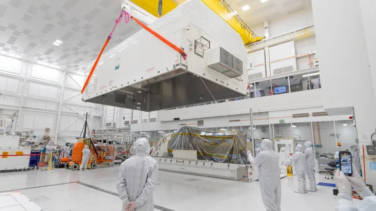 Technicians in a cleanroom oversee the crane lifting of a large white space payload container over a covered structure.