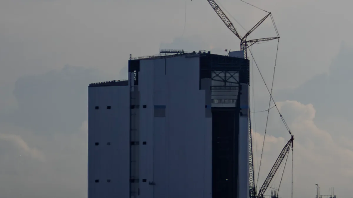 A large grey industrial assembly building under construction with a tall crane against a cloudy sky.