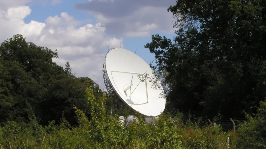 A large white satellite dish stands amidst dense green foliage under a partly cloudy sky.