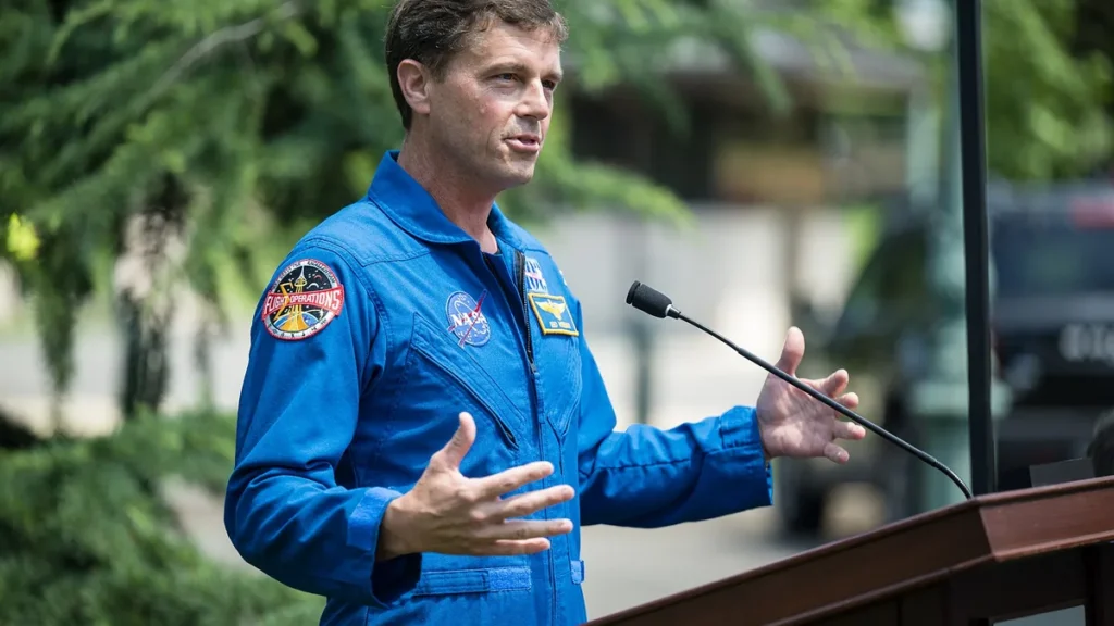 An astronaut in a blue NASA flight suit gestures while speaking at a podium.