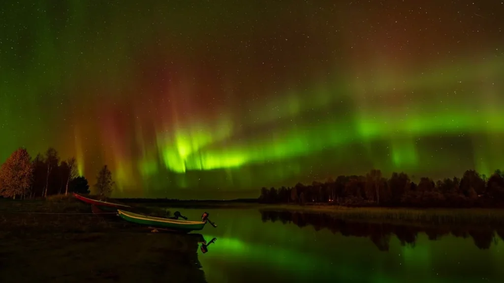 Green and red aurora borealis reflecting in a calm lake with a small boat on the shore.