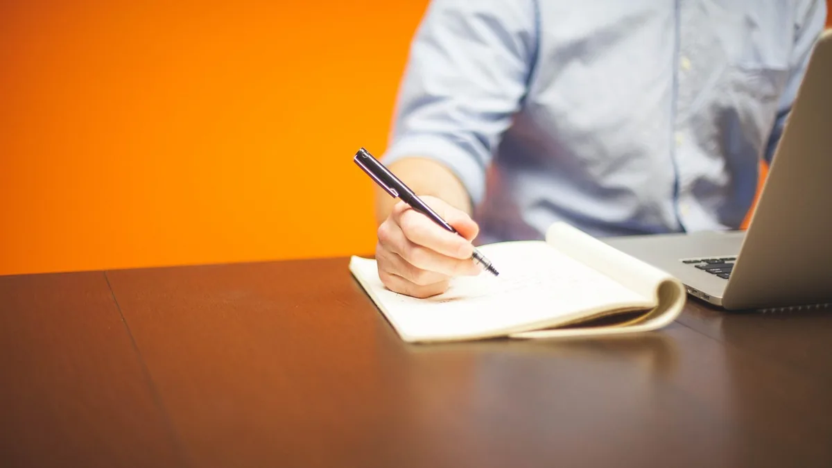 A person writing in a notebook with a pen next to a laptop on a wooden desk.