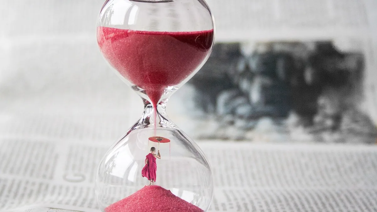 A miniature monk holding a red umbrella stands under a stream of red sand inside a glass hourglass.