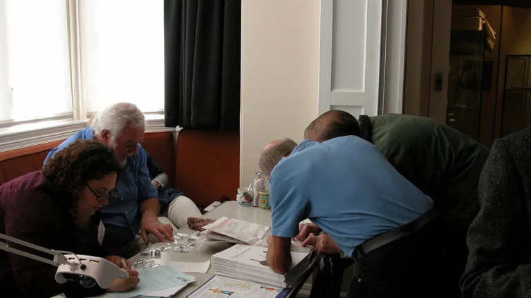 A group of people gathered around a table closely examining maps, documents, and small items under a lamp.