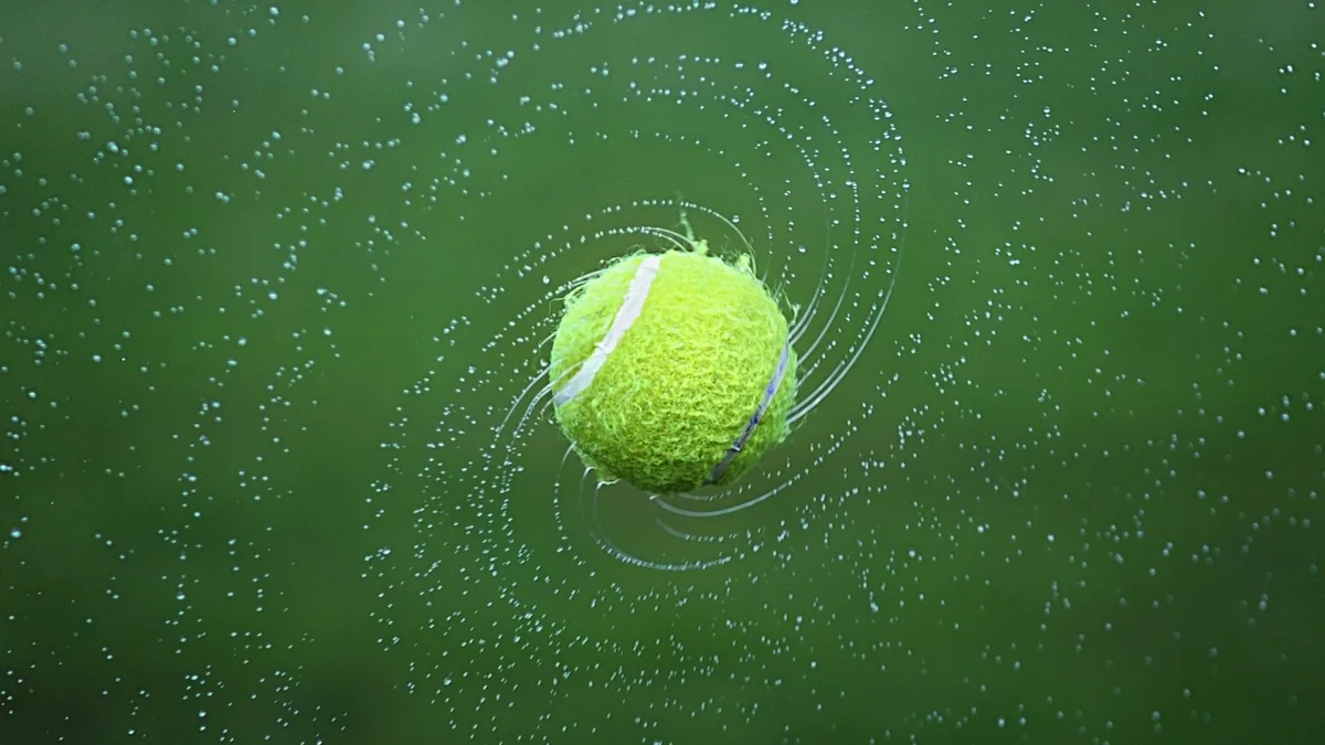 Green tennis ball spinning fast, surrounded by spiraling water droplets on a green background.