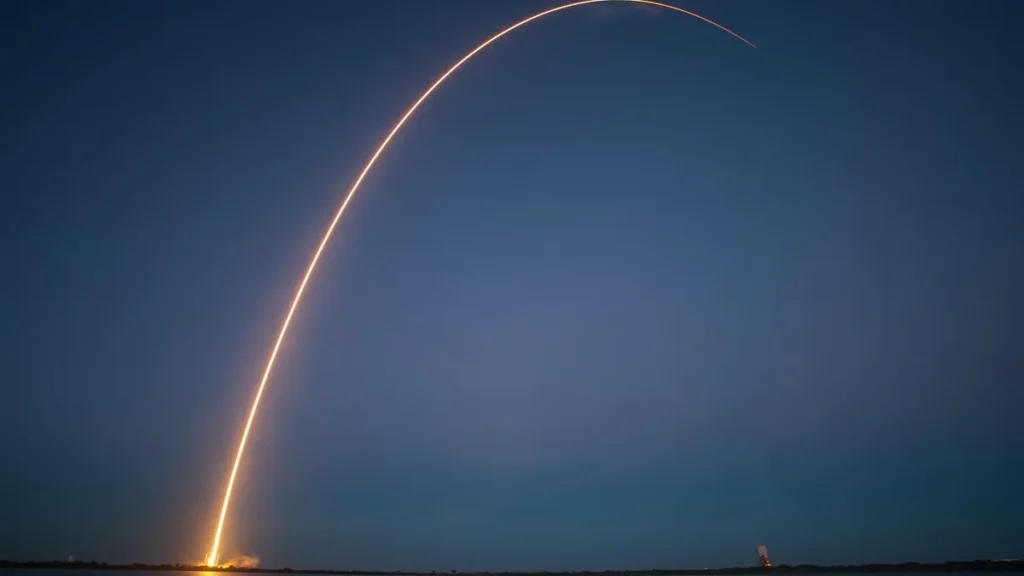 Bright orange rocket launch trail forming a high arc against a deep blue twilight sky.