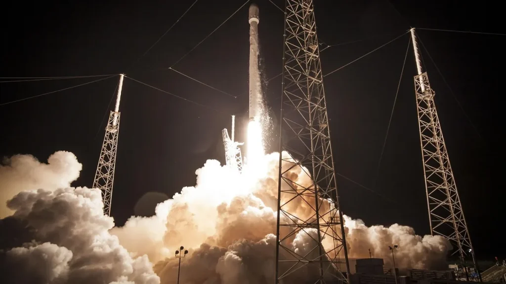 A rocket launches into the night, surrounded by a massive cloud of bright smoke and towering metal towers.