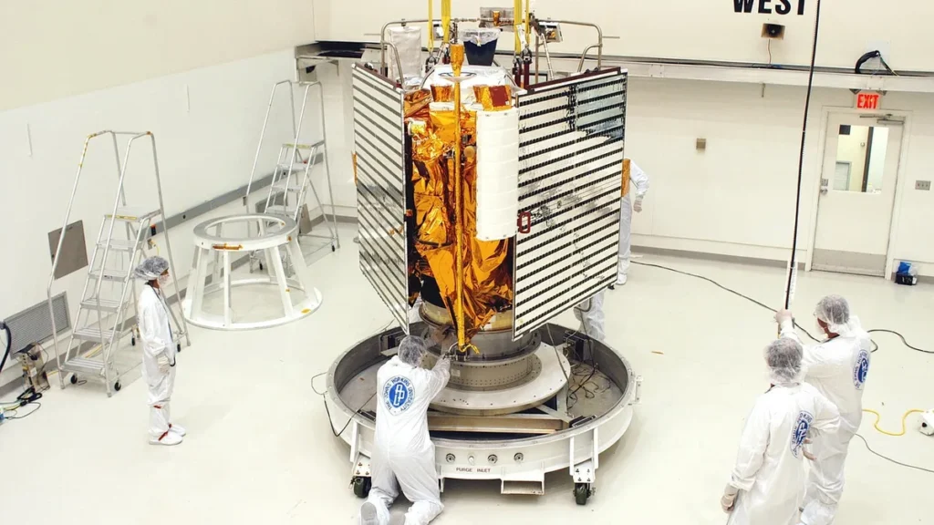 Technicians in cleanroom suits assemble a gold-wrapped satellite with deployed solar panels.
