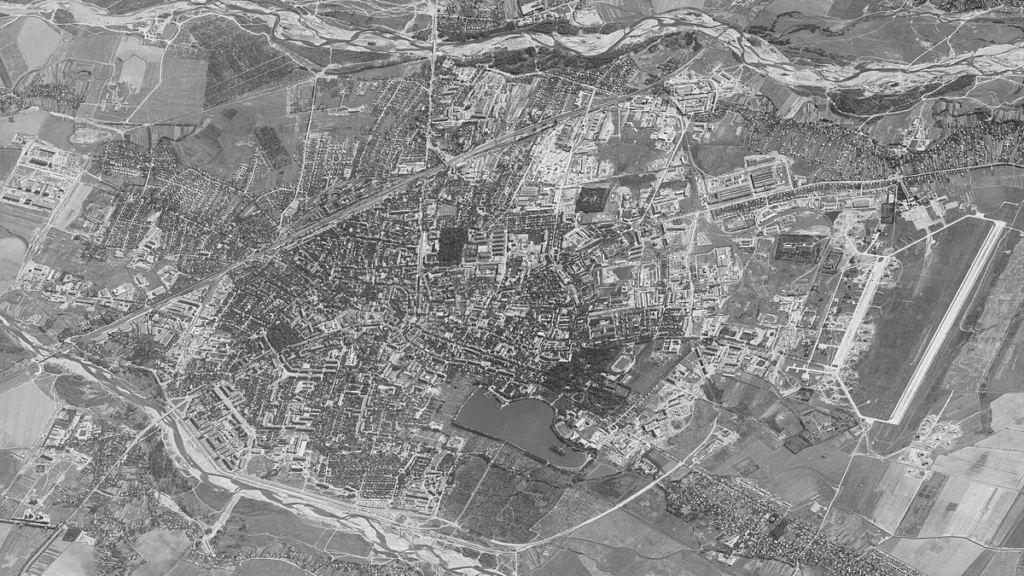 High-resolution black-and-white satellite imagery showing a city layout with a large airfield and surrounding rivers.