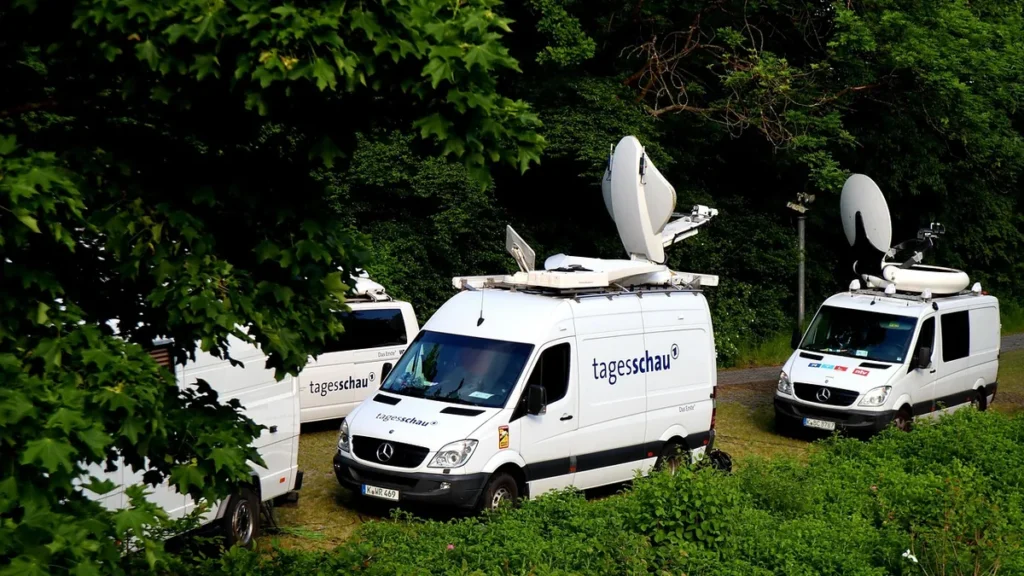 White Mercedes vans with tagesschau logos and large satellite dishes parked near green trees.