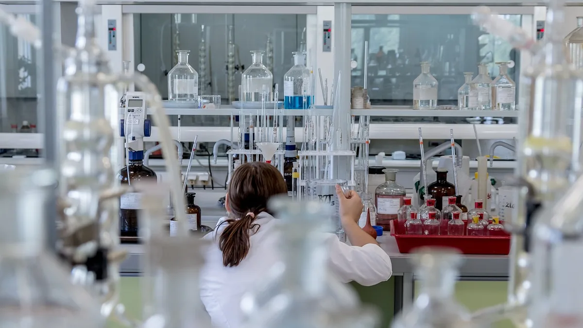 A scientist in a white coat works with glassware and bottles in a bright, busy laboratory.