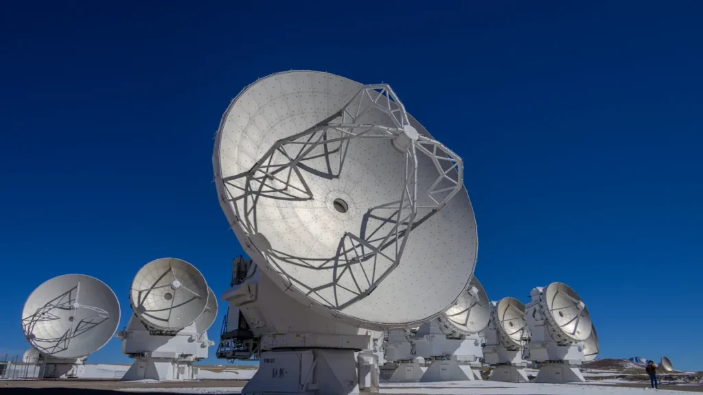 Large white radio telescope dishes point skyward in a high-altitude observatory array under a clear blue sky.