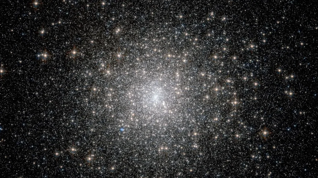 A dense, glittering cluster of thousands of stars against a dark cosmic background, with a bright blue star nearby.