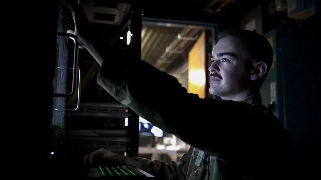 A military technician in a dark server room works on network hardware with a green-backlit keyboard.