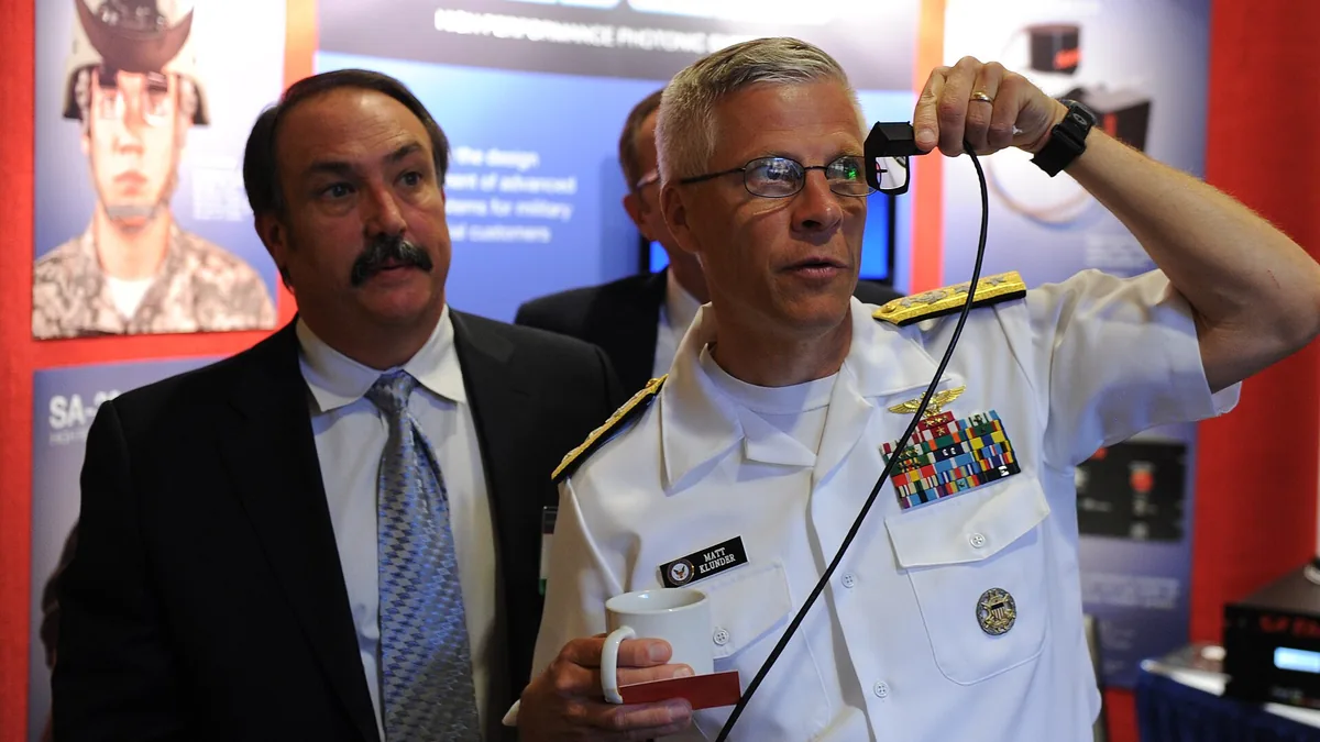 US Navy Admiral Matt Klunder holds a small black photonic device at a technology exhibition, with a colleague in a suit