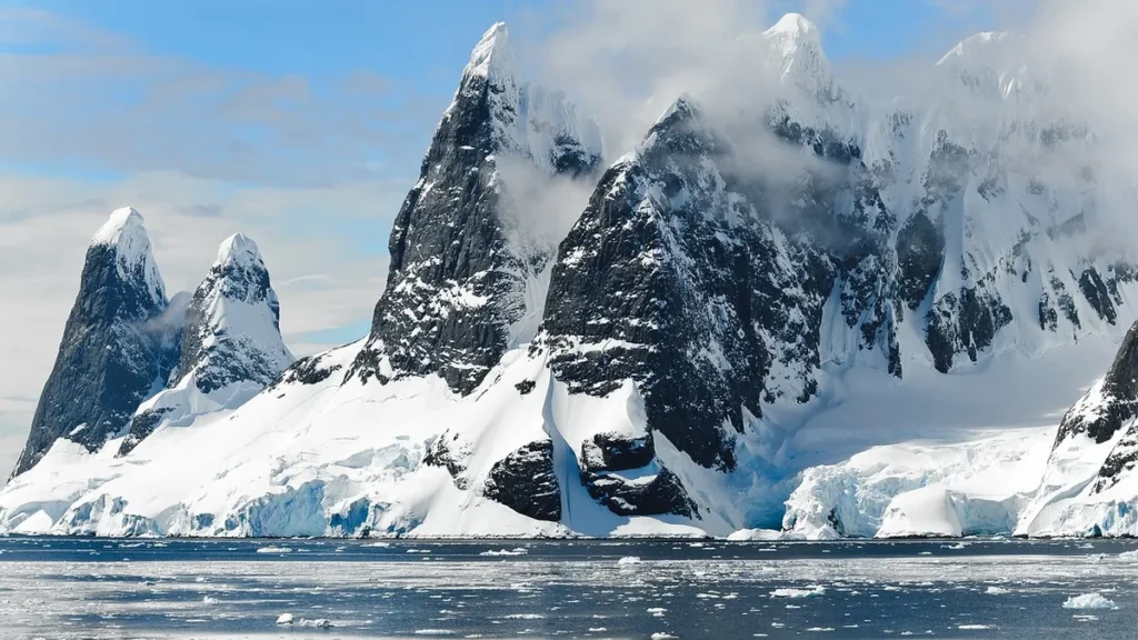 Snow-covered jagged peaks rise from icy Antarctic waters under a partly cloudy blue sky.