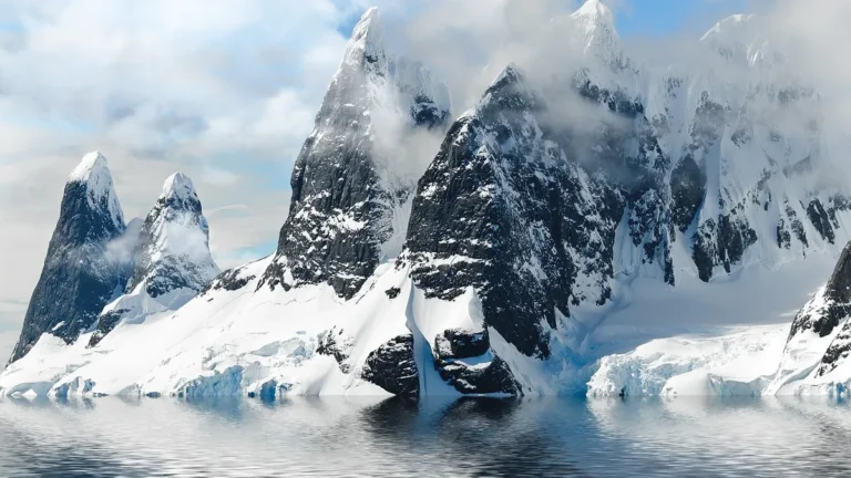 Snow-covered jagged peaks rise from the calm Antarctic waters, shrouded in mist and clouds.