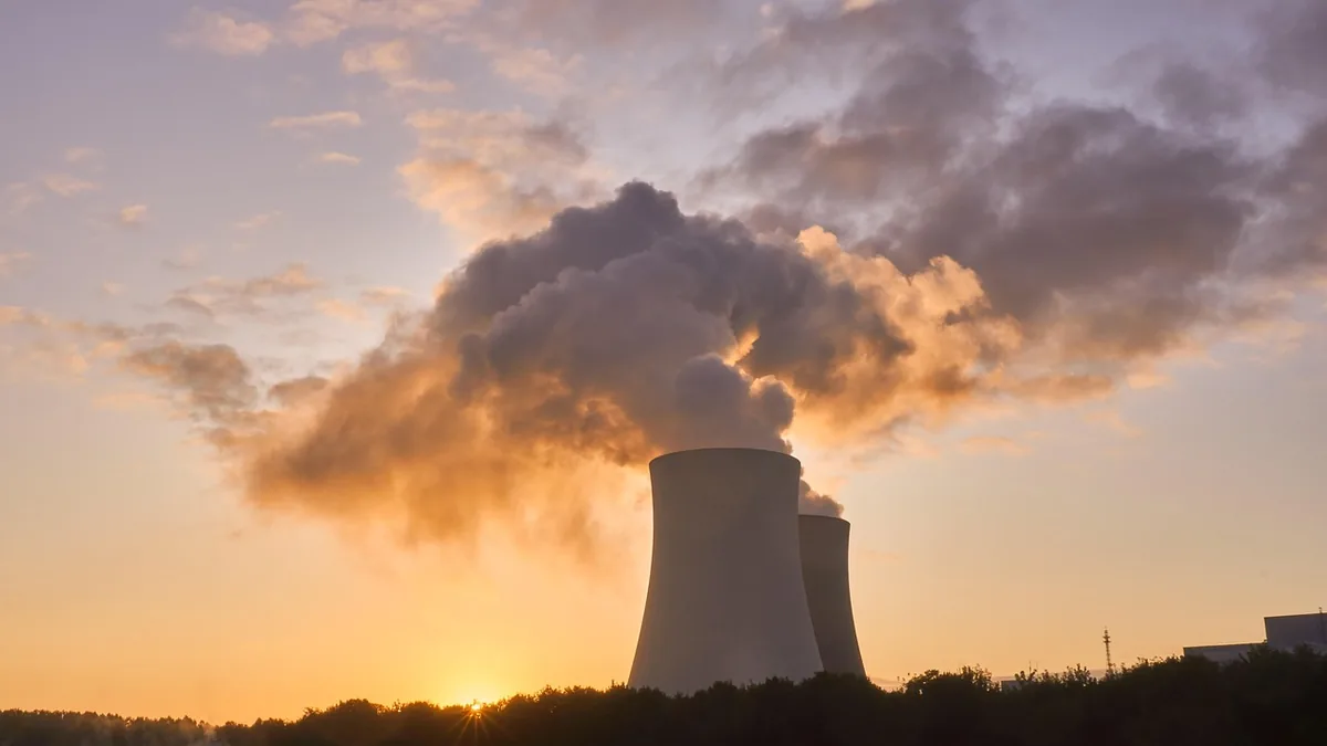 Two giant cooling towers releasing thick clouds of steam against a vibrant orange sunset sky.
