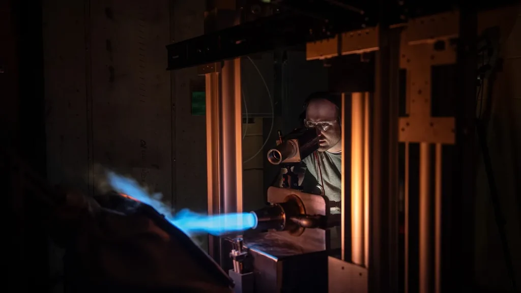 A researcher looks through optical equipment while monitoring a bright blue flame in a dark laboratory.