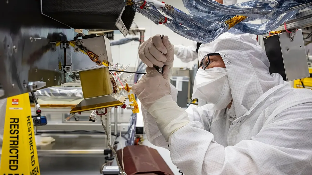 A technician in a white cleanroom suit and mask carefully works on gold-colored space hardware.