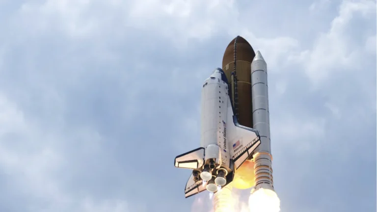 Space Shuttle Atlantis launches into a cloudy sky, showing bright fiery exhaust from its rocket boosters.