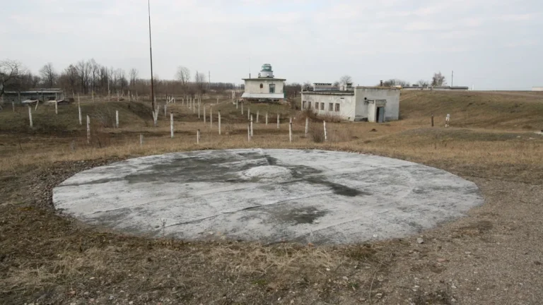 Concrete circular pad and old space tracking buildings in a dry grassy field.