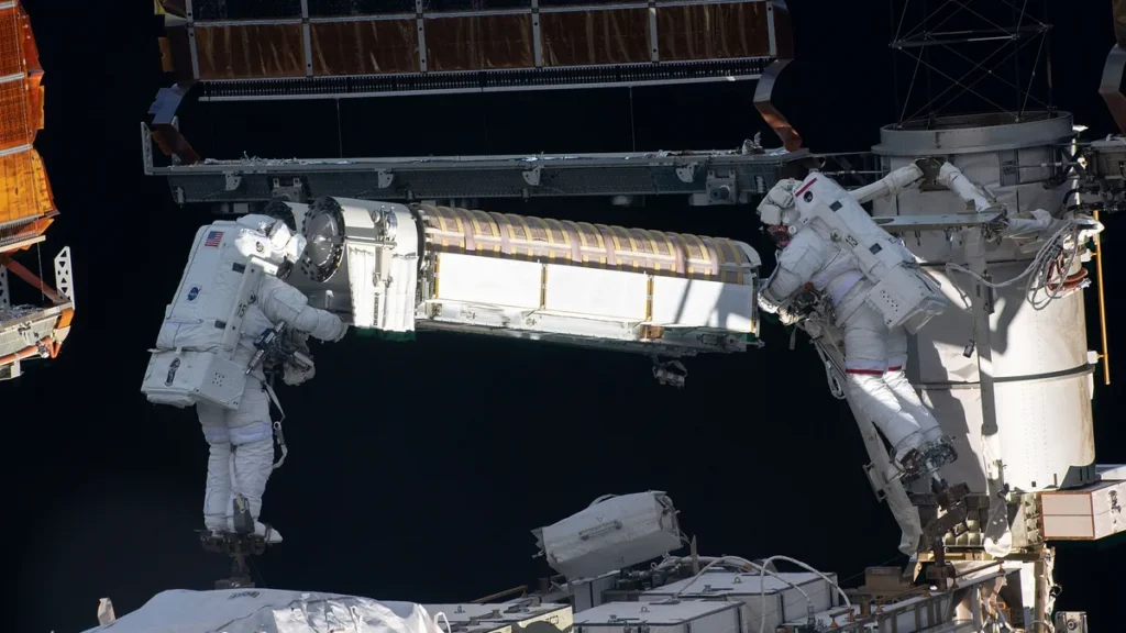 Two astronauts in spacesuits work on a large module during a spacewalk outside the ISS.