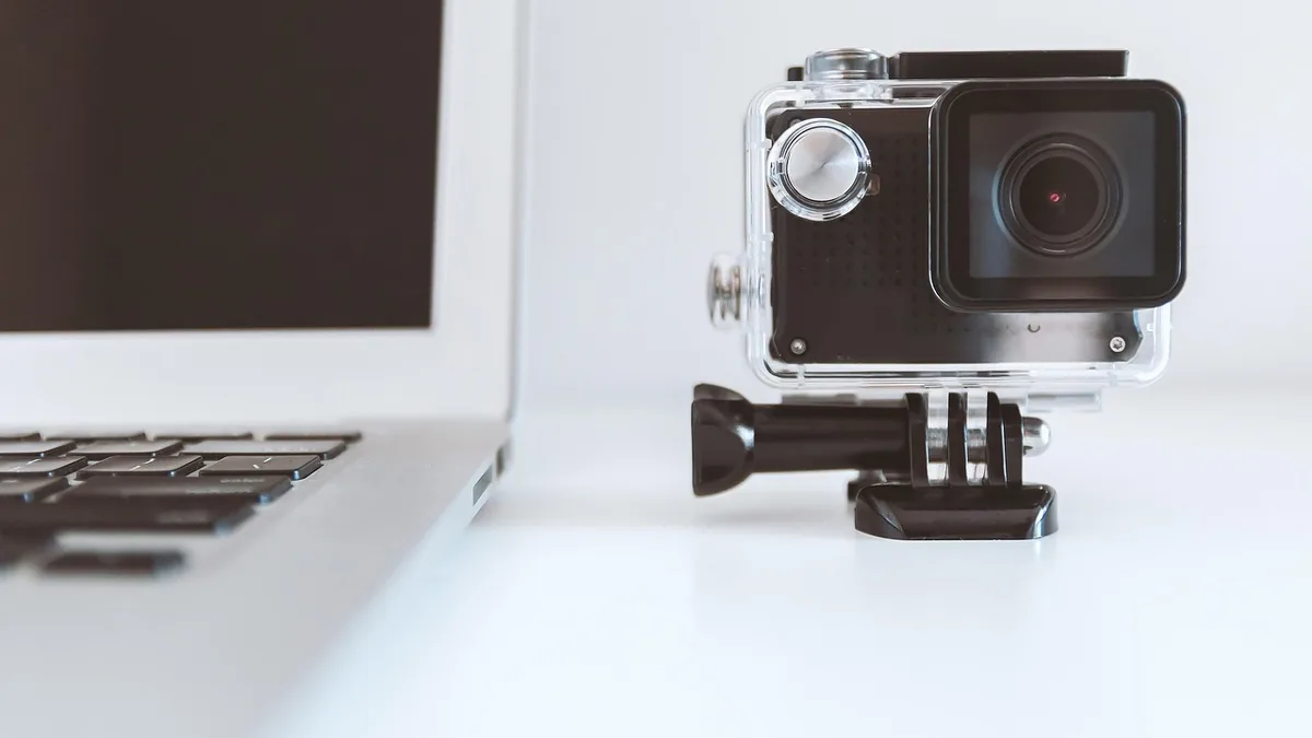 An action camera in a clear waterproof housing sits on a white desk next to a silver laptop keyboard.