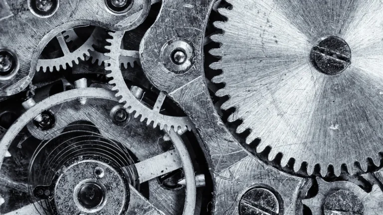 A black and white close-up of interlocking metallic gears, screws, and watch mechanism parts.