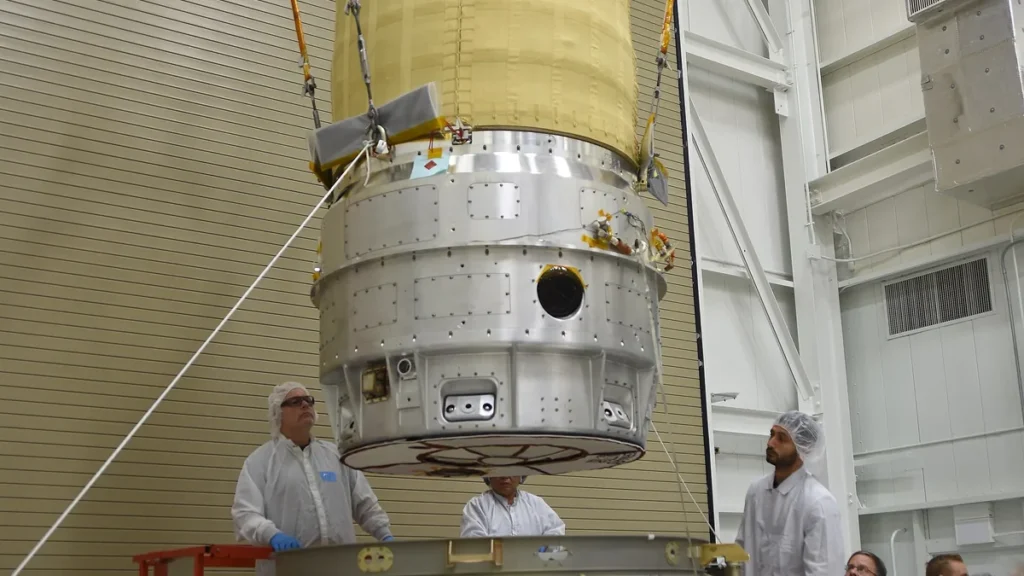 Technicians in cleanroom suits guide a large metallic spacecraft component suspended by crane cables.