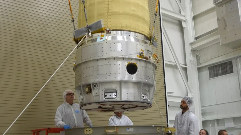 Technicians in cleanroom suits guide a large metallic spacecraft component suspended by crane cables.