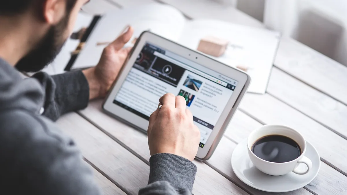 Person reading news on a white tablet with a cup of coffee on a wooden table.