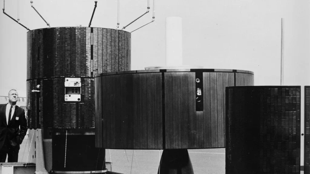 Archival photo of large cylindrical satellites covered in solar panels, with a man in a suit looking up on the left.