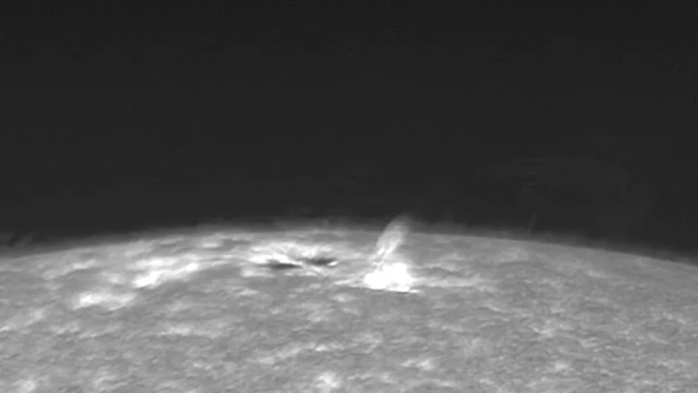 Black-and-white telescope view of a bright solar flare erupting from the Sun's textured surface.