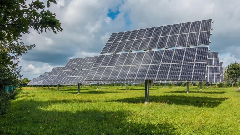 Rows of large solar panels stand in a green field under a partly cloudy sky.