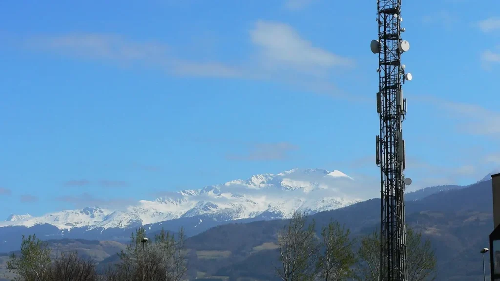 A tall telecommunications tower stands against a backdrop of snow-capped mountains and a blue sky.