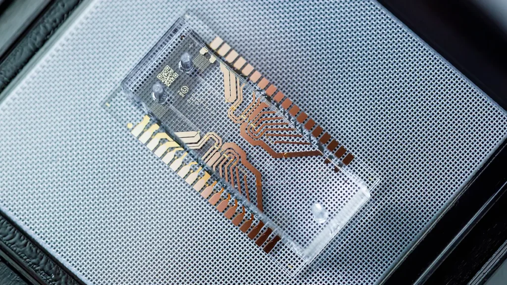 A transparent microfluidic chip with copper-colored circuit traces and a QR code on a grey grid.