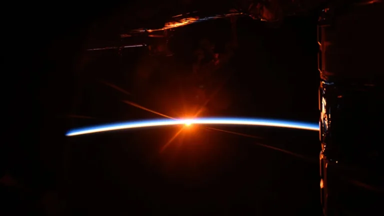 Vibrant orange sunrise over Earth's blue horizon, framed by the dark silhouette of an orbital spacecraft.