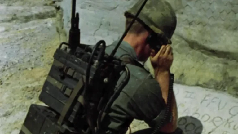 A soldier in a helmet and military uniform speaks into a receiver attached to a large backpack radio transmitter.