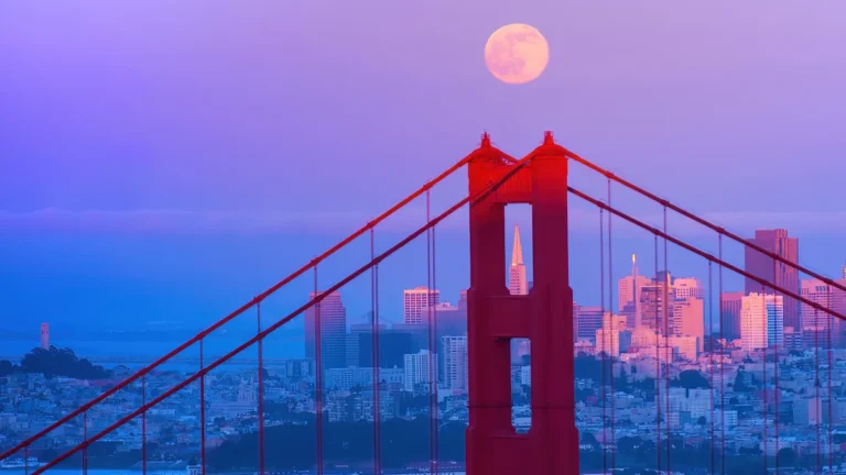 Full moon over the Golden Gate Bridge tower with the San Francisco skyline in purple twilight.