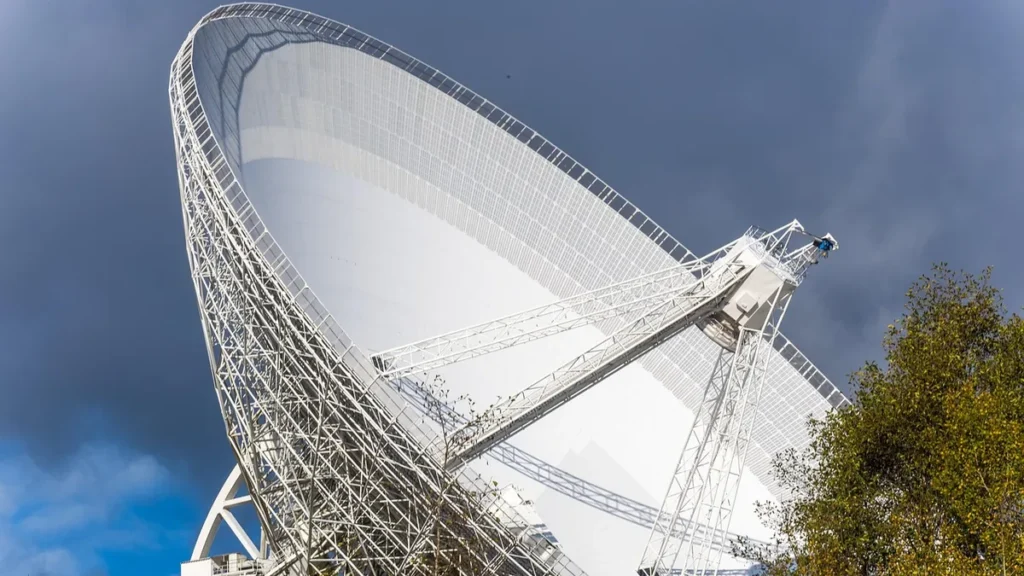 Large white radio telescope dish against a cloudy sky, partially framed by green trees.